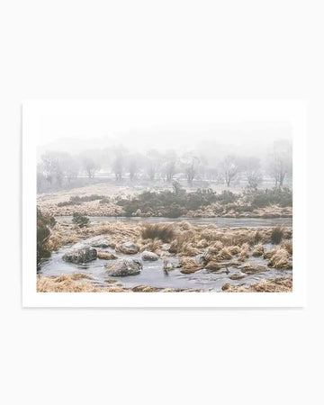 Framed photography print of a misty winter landscape featuring a frozen lake and frosted golden-brown grasses in the foreground, with a line of bare trees shrouded in fog stretching across the midground. The print has a natural wood frame and white border.