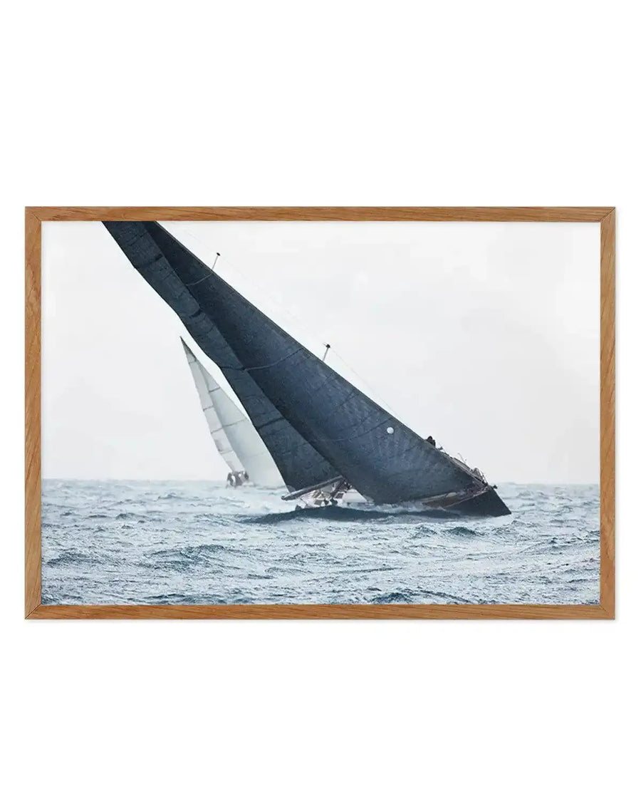 Framed photograph of two sailboats racing on choppy water under an overcast sky, with a natural wood frame and white border. The foreground features a dark grey sailboat leaning heavily into the wind, its sail taut, creating a wake. Behind it, a lighter grey sailboat is also visible, partially obscured. The water is a deep blue-grey with visible waves, and the sky is a soft, diffused white.