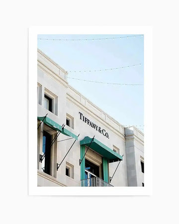 Architectural photograph printed on paper with white border, framed in white. The image shows a cream-colored historic building facade with a green awning displaying "TIFFANY & CO." in dark letters. Features symmetrical windows, ornate molding, decorative iron railings on a balcony beneath the awning, and string lights crossing the clear blue sky above. Captures the elegant storefront of the iconic luxury jewelry retailer located on Rodeo Drive.
