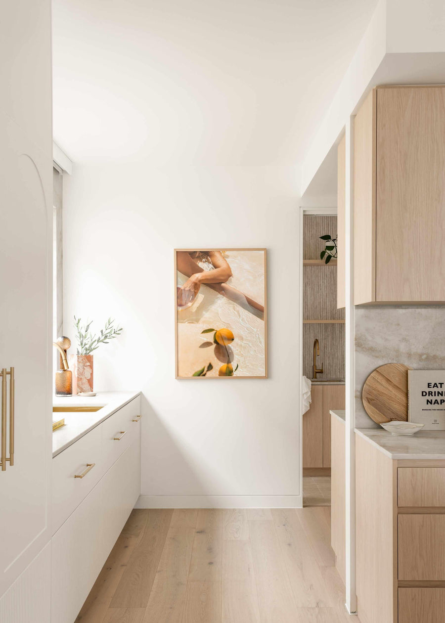 Lifestyle shot of a modern kitchen with a framed art print displayed on a white wall. The print, titled "Pool Time II," is a photograph featuring a person's legs partially submerged in clear, shallow water with two oranges floating nearby, creating a warm, summery mood. The frame is natural wood with a white border. The kitchen features light wood cabinetry on the right, white lower cabinets with gold handles on the left, and light wood flooring.