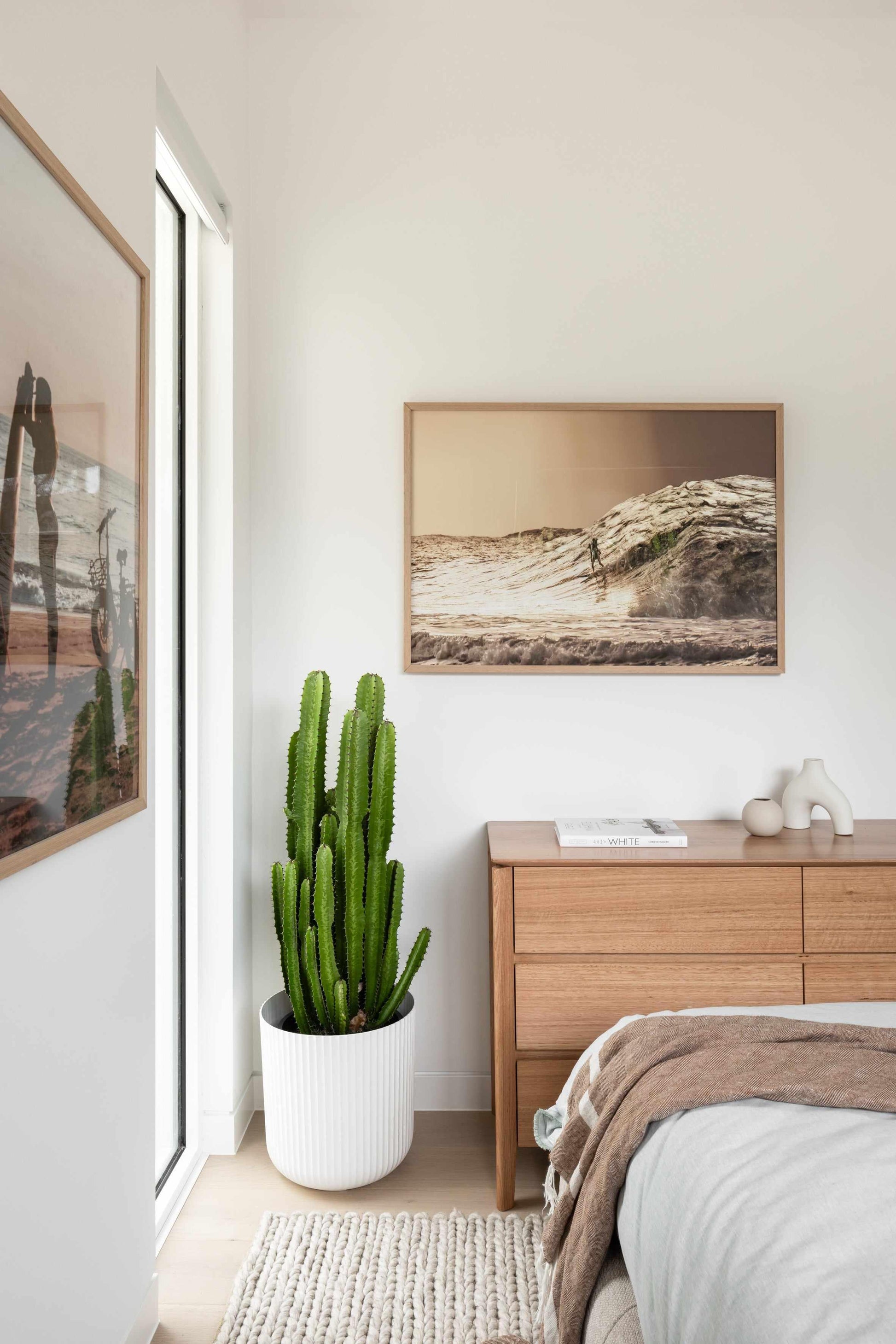 "Large framed art print displayed above a wooden dresser in a minimalist bedroom. The print features a sepia-toned photograph of a surfer riding a large wave at sunset, with a natural wood frame and no border. The room includes a tall green cactus in a white ribbed pot, a light wood dresser with two decorative vases and a book, a light grey bed with a brown throw blanket, and a cream textured rug."