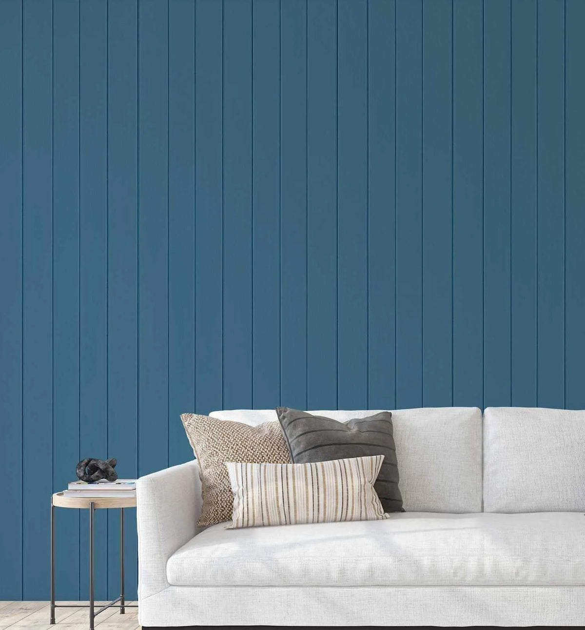 Lifestyle shot of a living room featuring a white sofa with neutral-toned cushions against a navy blue wall with vertical tongue and groove wood paneling. A small round side table with a black sculptural object and books sits to the left of the sofa.