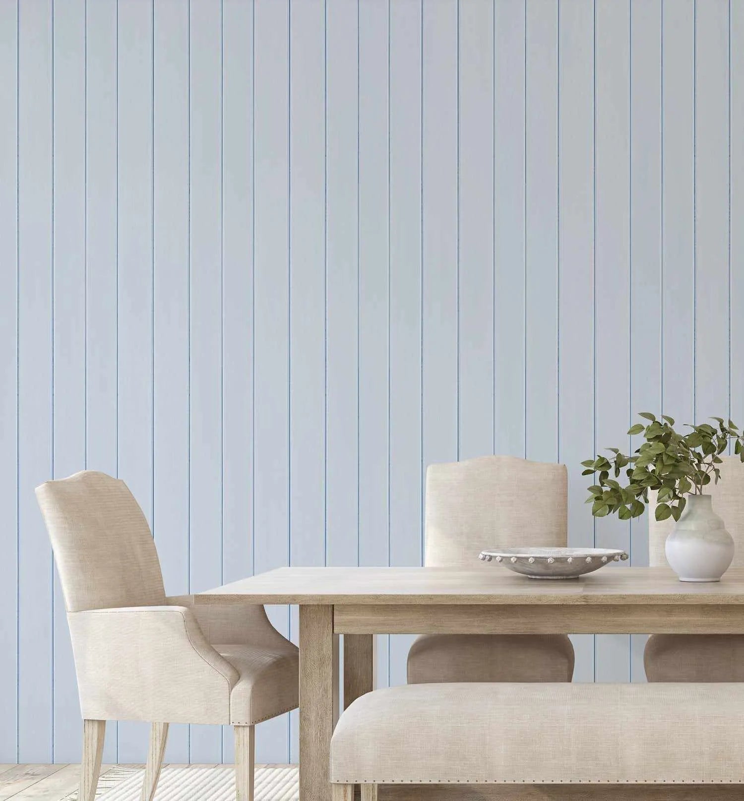 Tongue & Groove Wood Panel Wallpaper in Ice Blue displayed on a dining room wall behind a natural wood table set with cream upholstered chairs and a bench. The wallpaper features a vertical planked wood design in soft, pale blue-grey tones with subtle darker blue-grey grout lines between each panel, creating a realistic wood texture and finish. A white ceramic vase with green foliage and a decorative metal bowl sit on the table, demonstrating the wallpaper's calm, coastal aesthetic.