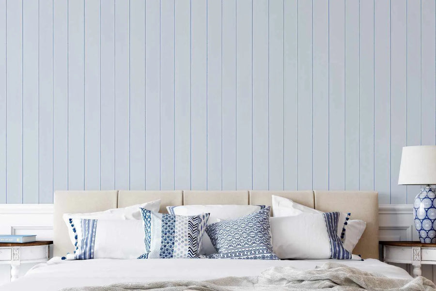 Bedroom wall displaying Tongue & Groove Wood Panel Wallpaper in ice blue, featuring evenly spaced vertical lines creating a realistic wood plank texture. The wallpaper has a soft, muted blue-gray tone with subtle depth. A cream-upholstered bed with blue and white patterned pillows and white bedding is positioned against the wallpapered wall. Matching nightstands with blue and white ceramic lamps flank the bed, demonstrating how the wallpaper complements coastal and contemporary bedroom styling.