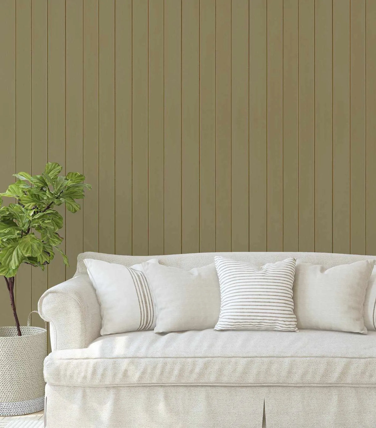 Tongue and Groove Wood Panel Wallpaper in khaki displayed on a living room wall behind a cream-colored sofa. The wallpaper features evenly-spaced vertical wood planks in a warm, muted sage-green tone with subtle texture and depth. The planks create a linear, beadboard-style pattern across the entire wall surface. A potted green plant sits to the left of the sofa, and the sofa is accessorized with five decorative pillows in cream, white, and sage striped fabrics.