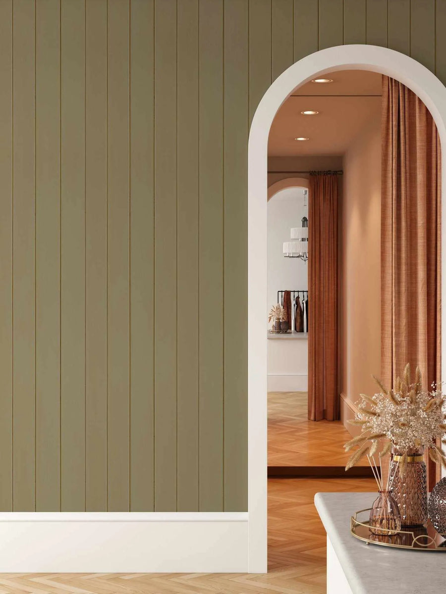 Lifestyle shot of a room featuring Tongue & Groove Wood Panel Wallpaper in Khaki. The wallpaper covers a wall with vertical panels in a muted, earthy khaki green, extending from a white baseboard to the ceiling. An arched doorway on the right reveals an interior space with warm lighting, wood flooring laid in a herringbone pattern, and rust-colored curtains. In the foreground, a white countertop holds a gold tray with decorative vases containing dried pampas grass and other dried botanicals.