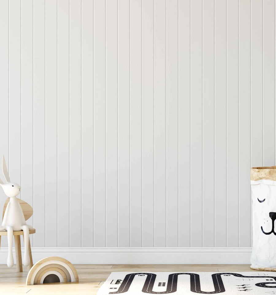Lifestyle shot of a child's room featuring Tongue & Groove Wood Panel Wallpaper in Light Grey. The wallpaper covers a wall with vertical panels and a white baseboard. A white bunny toy sits on a small wooden chair on the left, and a wooden rainbow toy is on the floor. A white rug with a black road pattern and a white bear-shaped storage basket are on the right.