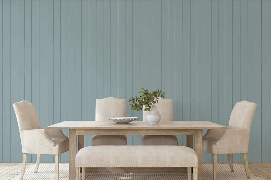 Dining room interior featuring Tongue & Groove Wood Panel Wallpaper in soft sage green with vertical wood plank texture. The wallpaper covers the full wall behind a light natural wood dining table set with cream upholstered chairs and a matching bench. A white ceramic vase with green foliage and decorative bowl sit on the table. The wallpaper's subtle vertical striping creates a calm, sophisticated backdrop with a matte finish that mimics real wooden paneling in a muted blue-green tone.