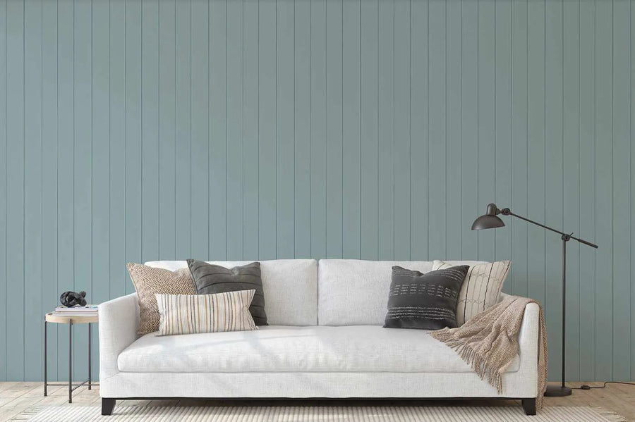 Lifestyle shot of a living room featuring a white sofa with various throw pillows and a floor lamp, set against a wall covered in Tongue & Groove Wood Panel Wallpaper in a soft blue-green hue. The wallpaper features vertical panels with subtle grooved details, creating a classic, textured wainscoting effect.