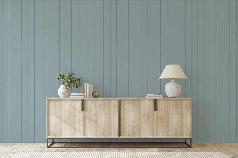 Tongue and groove wood panel wallpaper in a soft, muted blue-green hue, creating a serene backdrop in a modern living room. The wallpaper features vertical panels with subtle shadows, adding depth and texture to the wall. A light wood sideboard with dark metal handles is positioned against the wall, adorned with a white textured vase holding green foliage, stacks of books, decorative spheres, and a white ceramic lamp with a cream shade. The room has light wood flooring and a cream striped rug.