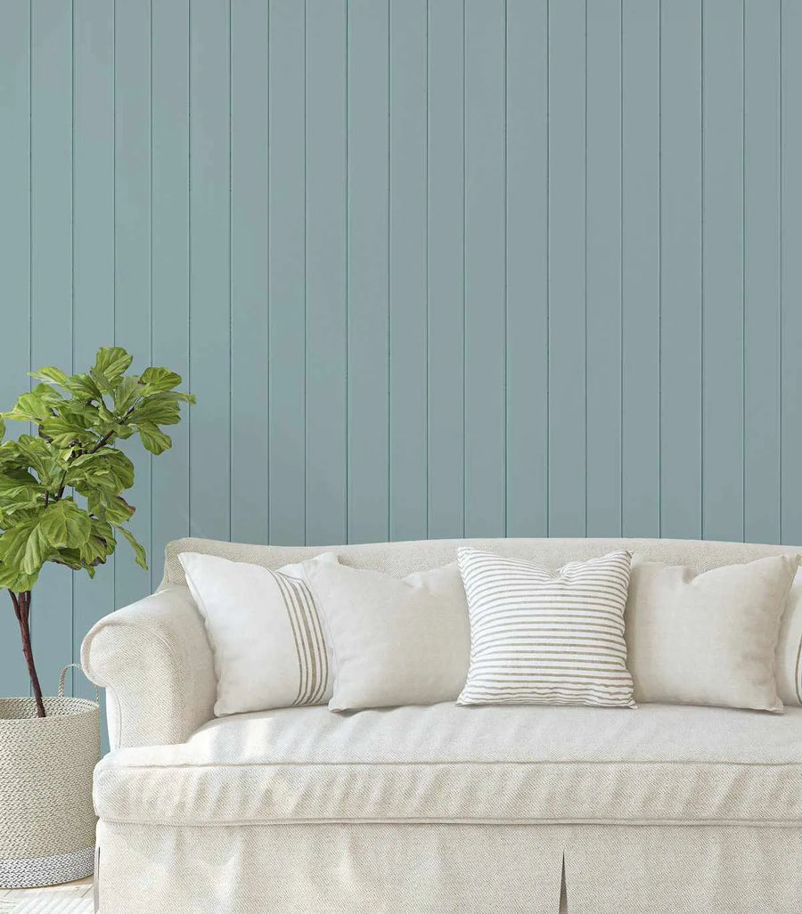 Lifestyle shot of a living room featuring Tongue & Groove Wood Panel Wallpaper in Mountain Rivers. The wallpaper is a soft, muted blue-green with vertical lines mimicking wood panels, creating a serene backdrop. A cream-colored sofa with a slipcover is positioned in front of the wallpaper, adorned with several throw pillows in cream and white with subtle striped patterns. A large green plant in a woven basket sits to the left of the sofa.
