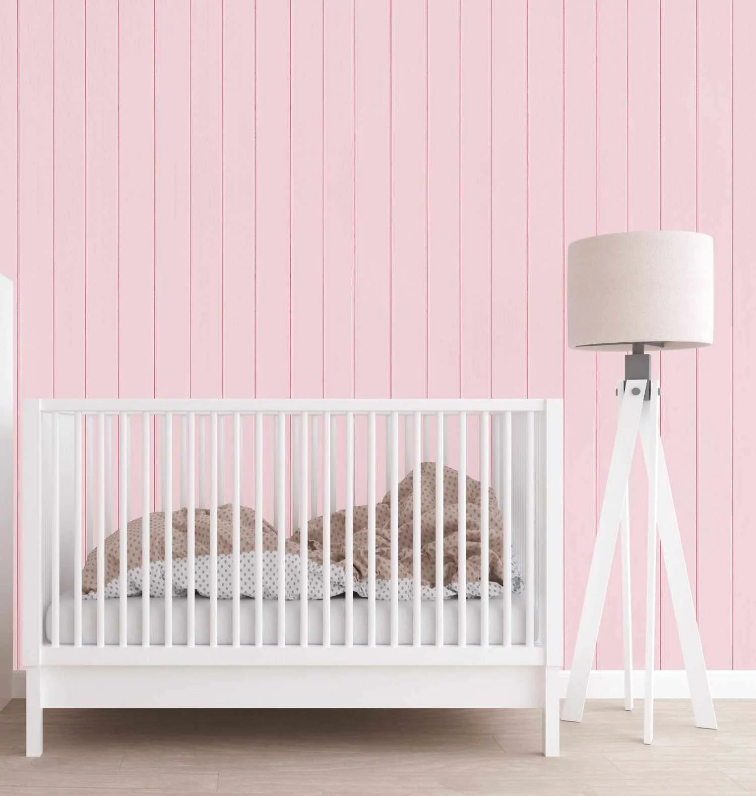 Nursery room with soft pink vertically paneled wood wallpaper covering the wall. A white wooden crib with natural wood bedding sits against the wall, accompanied by a white tripod floor lamp with cream linen drum shade. The wallpaper features evenly spaced vertical boards in a pale pink tone with subtle wood grain texture, creating a clean, minimalist aesthetic suitable for a baby's room.