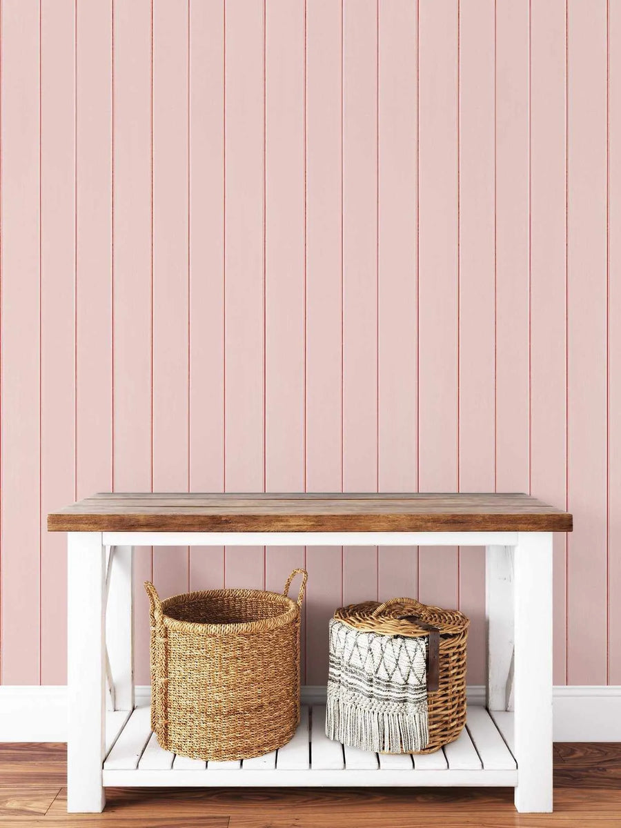 Tongue & Groove Wood Panel Wallpaper in dusty rose displayed on a wall behind a white wooden shelf with natural wood top. The wallpaper features evenly spaced vertical planks in a soft mauve-pink tone with subtle brown grout lines, creating a realistic shiplap effect. Two woven baskets in natural tan and golden tones sit on the shelf against the wallpaper background, demonstrating the soft, understated aesthetic of the design.