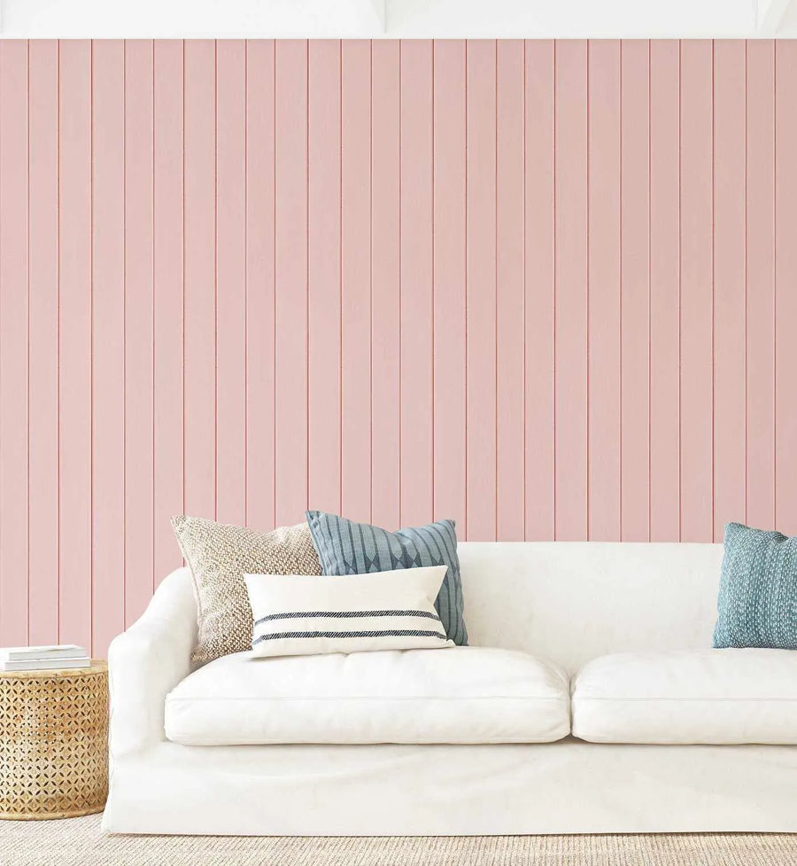 Lifestyle shot of a living room featuring a pink tongue and groove wood panel wallpaper behind a white slipcover sofa. The wallpaper has vertical panels in a soft blush pink with subtle reddish-brown lines indicating the grooves. The sofa is adorned with various pillows: a textured cream pillow, a blue and white striped pillow, and a solid light blue pillow. A round, woven wooden side table with books sits to the left of the sofa, and a textured cream rug covers the floor.