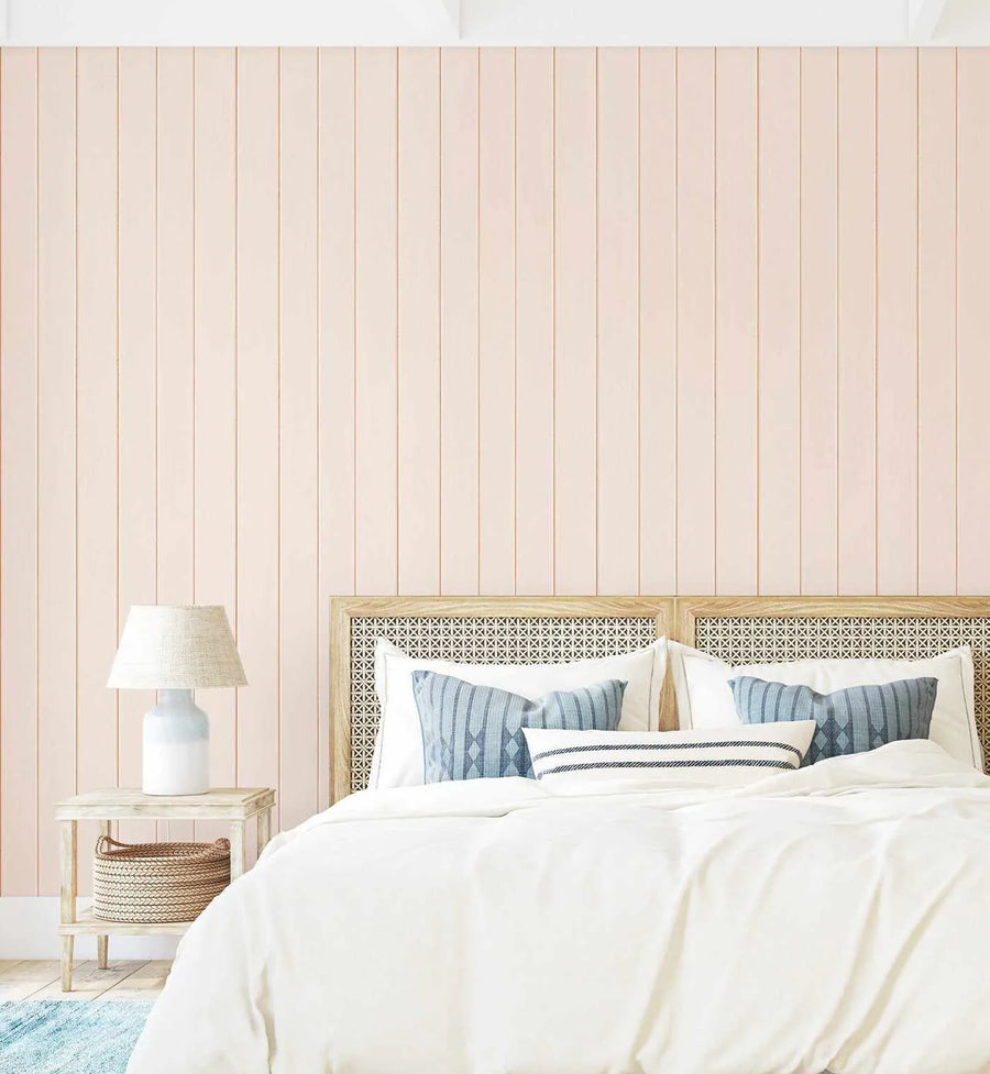 Bedroom wall displaying Tongue & Groove Wood Panel Wallpaper in a soft peachy-pink tone with subtle cream and taupe vertical wood grain lines creating a paneled effect. The wallpaper covers the wall behind a bed with a geometric patterned headboard, white bedding, and blue-striped decorative pillows. A wooden nightstand with a woven basket and white ceramic table lamp sits to the left. The overall aesthetic is coastal and minimalist with a warm, neutral color palette.