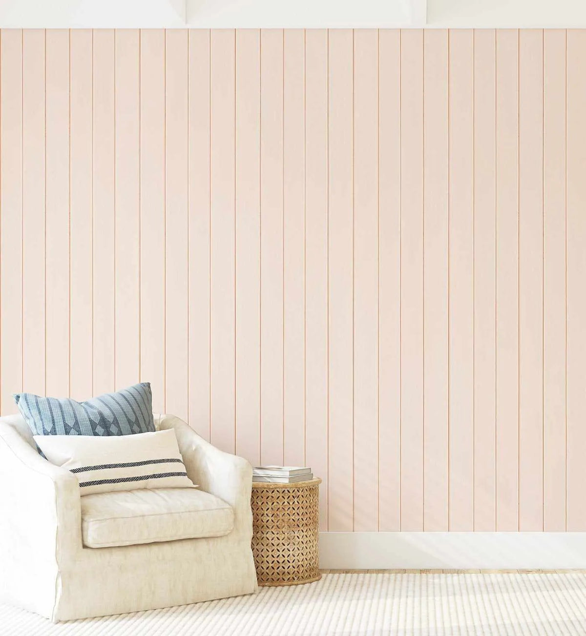 Tongue & Groove Wood Panel Wallpaper in Slightly Peach displayed on a bedroom wall. The wallpaper features a vertical wooden plank pattern in soft peachy-pink tones with subtle gray dividing lines creating individual planks. The wall extends from floor to ceiling, shown in a minimalist room setting with a cream linen armchair, striped throw pillows in blue and cream, a woven cylindrical side table, and light neutral carpeting. The finish has a soft, matte appearance resembling natural wood texture.