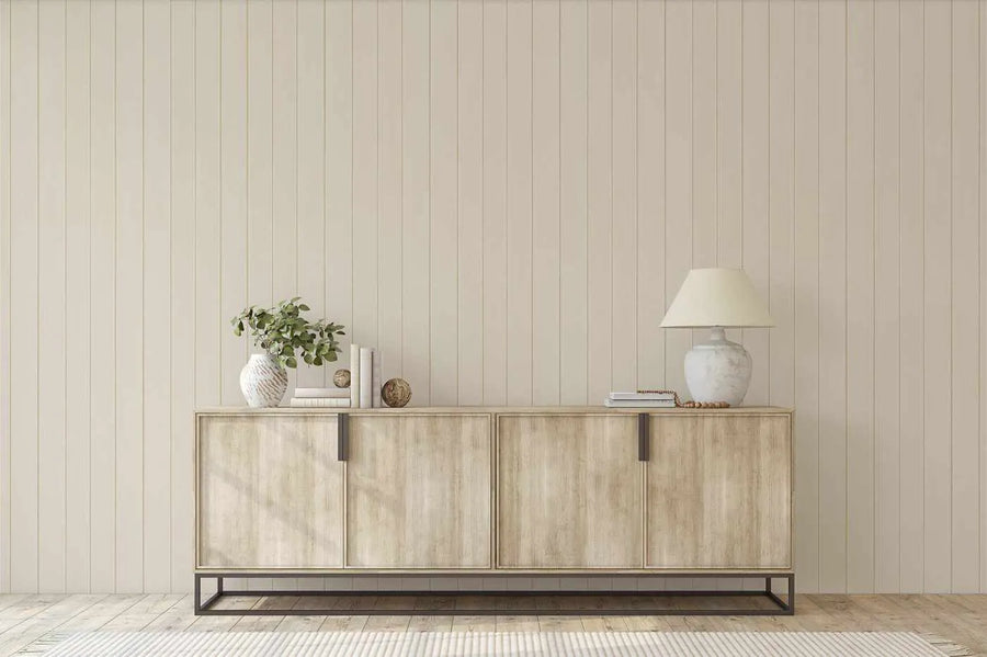 "Taupe tongue and groove wood panel wallpaper covering a wall behind a light wood sideboard in a minimalist living room. The wallpaper features vertical panels in a warm, muted taupe shade, creating a subtle textured backdrop. The sideboard is adorned with a white textured vase holding green foliage, a stack of books, decorative spheres, and a white ceramic lamp with a cream shade."