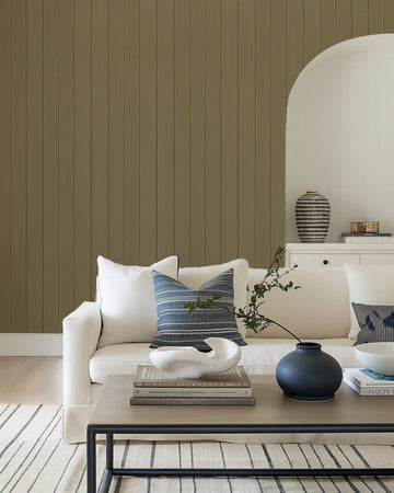 Khaki tongue and groove wood panel wallpaper displayed on a living room wall. The wallpaper features evenly spaced vertical planks in a warm, muted sage-green tone with subtle texture suggesting wood grain. The room setting shows a cream-colored sofa with blue and gray striped pillows, a white side table with decorative objects, and a light wood floor with a geometric patterned area rug, demonstrating how the wallpaper complements neutral, contemporary interior design.