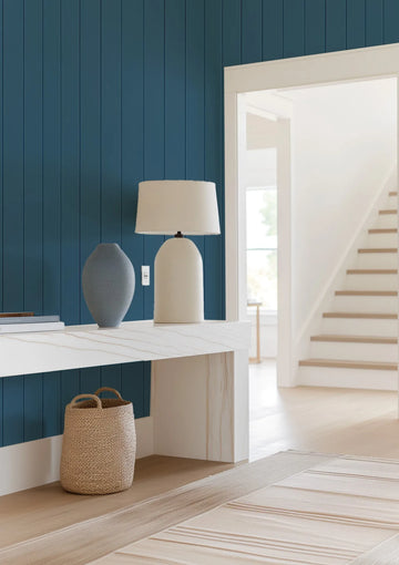 Lifestyle shot of a hallway with a navy blue tongue and groove wood panel wallpaper. The wallpaper covers the wall behind a white marble console table, which holds a grey vase and a cream-colored table lamp. A woven basket sits on the floor beneath the table. To the right, a doorway opens to a bright white room with a wooden staircase and light wood floors.