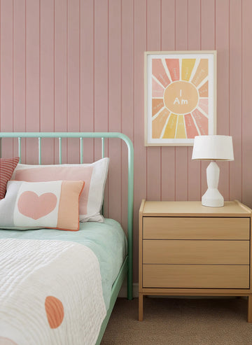 Lifestyle shot of a bedroom featuring a wall covered in peel and stick wallpaper with a repeating tongue and groove wood panel design in a soft, dusty rose pink. The wallpaper serves as a backdrop for a mint green metal bed frame with white and pink pillows, and a light wood bedside table with a white lamp. Above the bedside table, a framed art print with a sunburst design and the words "I Am" is displayed, featuring various positive affirmations in shades of orange, yellow, and pink.