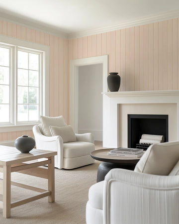 Lifestyle shot of a living room with tongue and groove wallpaper in a soft peach hue. The wallpaper features vertical panels with subtle grooved lines, creating a classic, textured look. The room includes a large window with white trim, a white fireplace with a black insert, and two cream-colored upholstered armchairs. A light wood coffee table and a round dark wood side table with books complete the inviting and minimalist space.