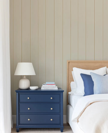 "A bedroom with a taupe tongue and groove wood panel wallpaper on the wall behind a bed with a woven headboard. A navy blue bedside table with gold knobs and a white lamp sits to the left of the bed. The bed is dressed in white linens with blue accent pillows. The wallpaper features vertical panels in a warm, muted taupe colour with subtle texture."