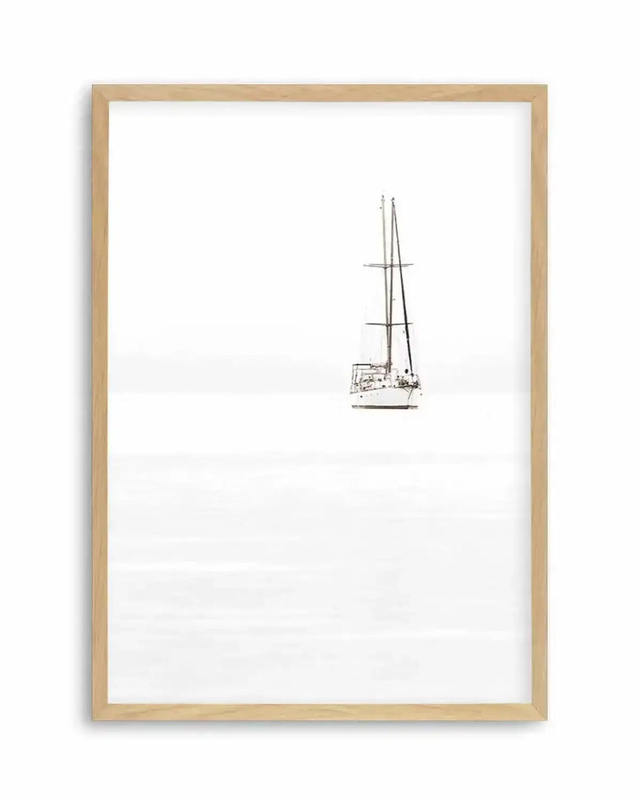 Framed black and white photograph of a sailboat at sea with a natural wood frame and white border. The composition features a single sailboat with two masts in the center, surrounded by a vast expanse of white, suggesting a foggy or minimalist ocean scene.