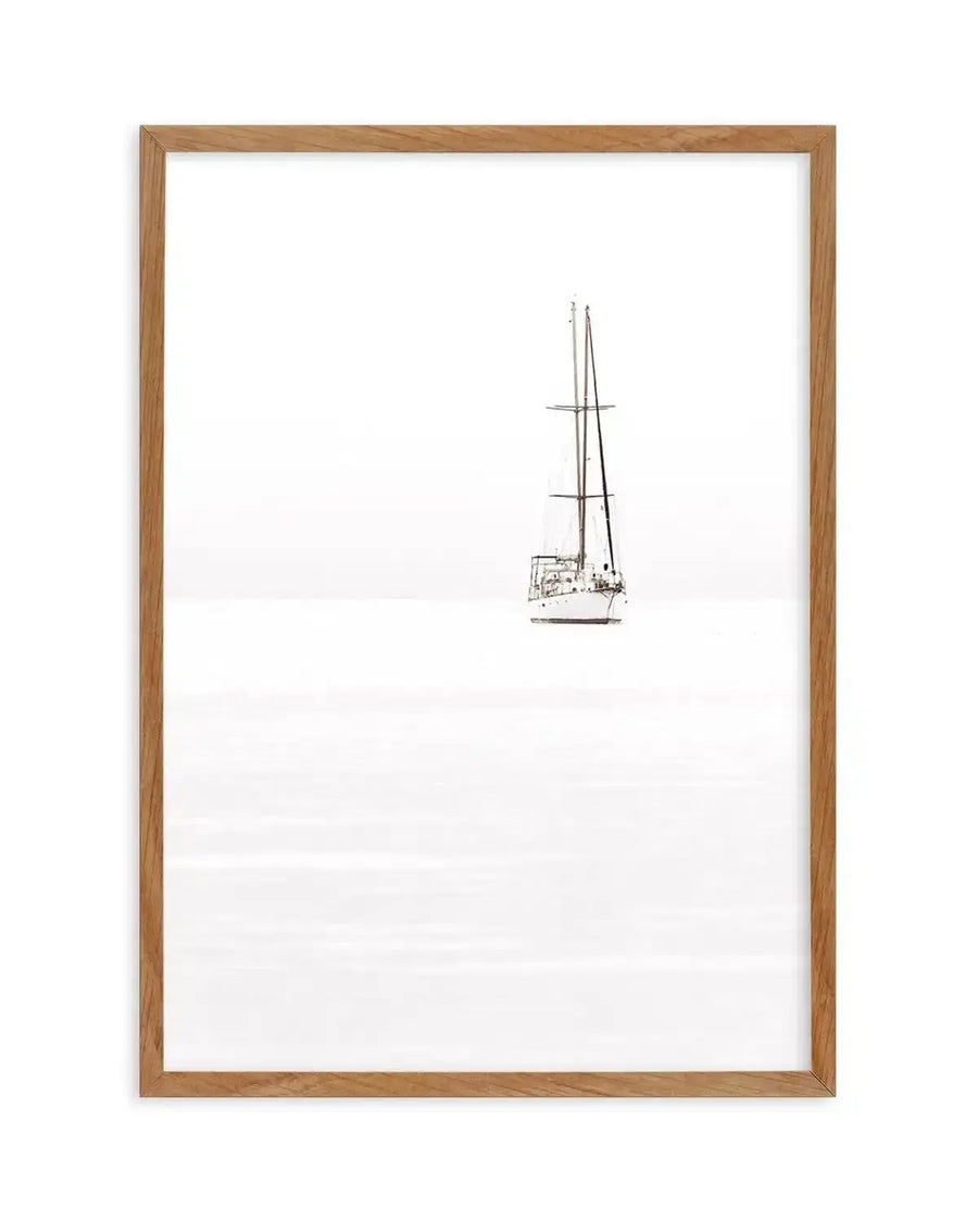 Framed minimalist photograph of a sailboat on a calm, bright white sea with a natural wood frame and white border. The sailboat, positioned slightly to the right of center, features two tall masts and a dark hull, appearing almost silhouetted against the stark white background.