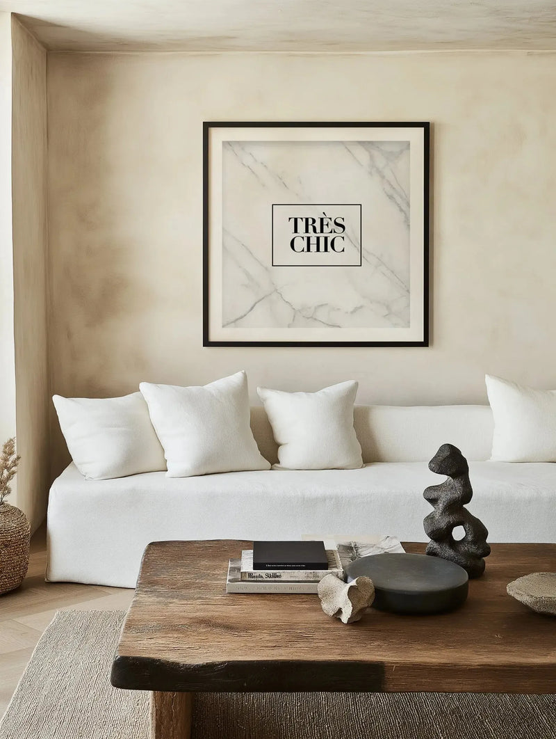 Framed art print displayed on a beige wall above a white upholstered sofa. The print features a marble-textured background in soft gray and white tones with subtle veining. Black text reading "TRÈS CHIC" is centered in a thin black rectangular border. The print is set in a black frame with a white border. The room setting shows minimalist décor with a wooden table, neutral textiles, and decorative objects in natural materials.