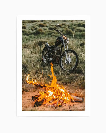 "Framed photograph of a vintage black motorcycle parked in a grassy field with a bonfire burning in the foreground, with white border. The motorcycle has chrome accents and a dark fuel tank, while the fire illuminates the sandy ground and dry grass around it under an overcast sky."