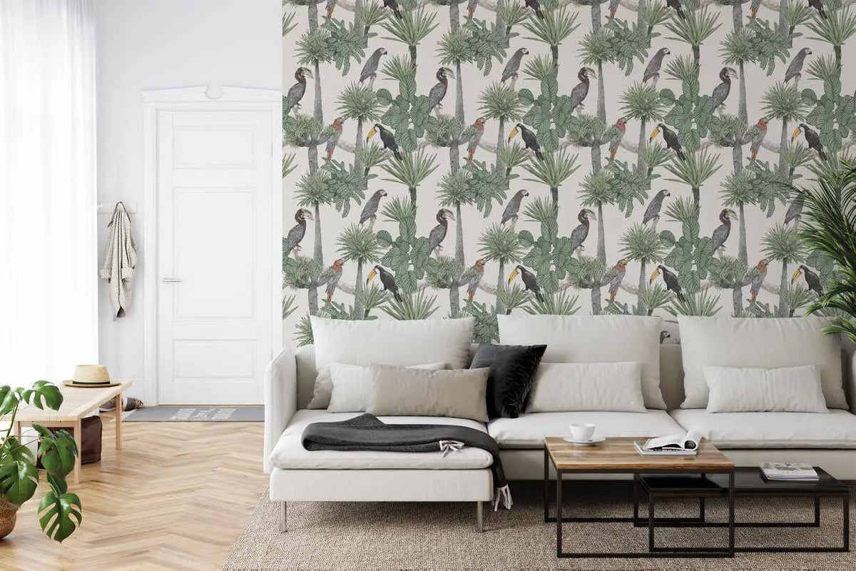 Peel and stick wallpaper mural featuring a repeating pattern of tropical birds and palm foliage in muted green, grey, and earth tones on a light cream background. The design includes illustrated toucans with distinctive beaked profiles and various palm fronds rendered in a naturalistic, botanical style. Displayed in a contemporary living room on an accent wall behind a cream-colored sectional sofa with grey and black accent pillows, wooden coffee table, and light wood herringbone flooring.