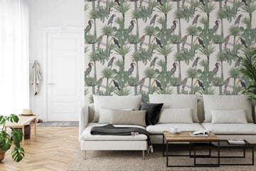 Peel and stick wallpaper mural featuring a repeating pattern of tropical birds and palm foliage in muted green, grey, and earth tones on a light cream background. The design includes illustrated toucans with distinctive beaked profiles and various palm fronds rendered in a naturalistic, botanical style. Displayed in a contemporary living room on an accent wall behind a cream-colored sectional sofa with grey and black accent pillows, wooden coffee table, and light wood herringbone flooring.