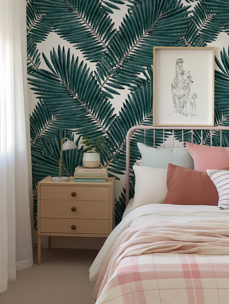 "Tropicana wallpaper featuring a repeating pattern of large-scale palm fronds in deep emerald green with visible brushstroke texture on a soft white background, displayed in a child's bedroom. The wallpaper is behind a pink metal bed frame with a light wood bedside table, a small plant, and a framed illustration of a child riding a lion."