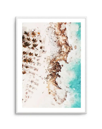 Framed aerial photograph of a tropical beach scene with a white frame and white border. The composition features a white sandy beach with scattered palm trees and tiny figures of people sunbathing and swimming, next to a rocky coastline, with clear turquoise ocean water on the right.