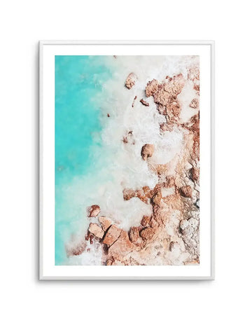 Framed aerial photography art print of a tropical beach scene with a white frame and white border. The composition features vibrant turquoise ocean water meeting a white sandy shore dotted with large, textured reddish-brown rocks, with white foamy waves breaking around them.