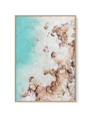 Aerial photograph framed in natural wood showing turquoise ocean water on the left transitioning to white foamy waves and sandy beach with tan and brown rocky formations on the right. The composition captures a coastal landscape from above with soft, hazy lighting and subtle color gradations from bright cyan water to cream and beige sand and stone. Natural wood frame with clean lines.