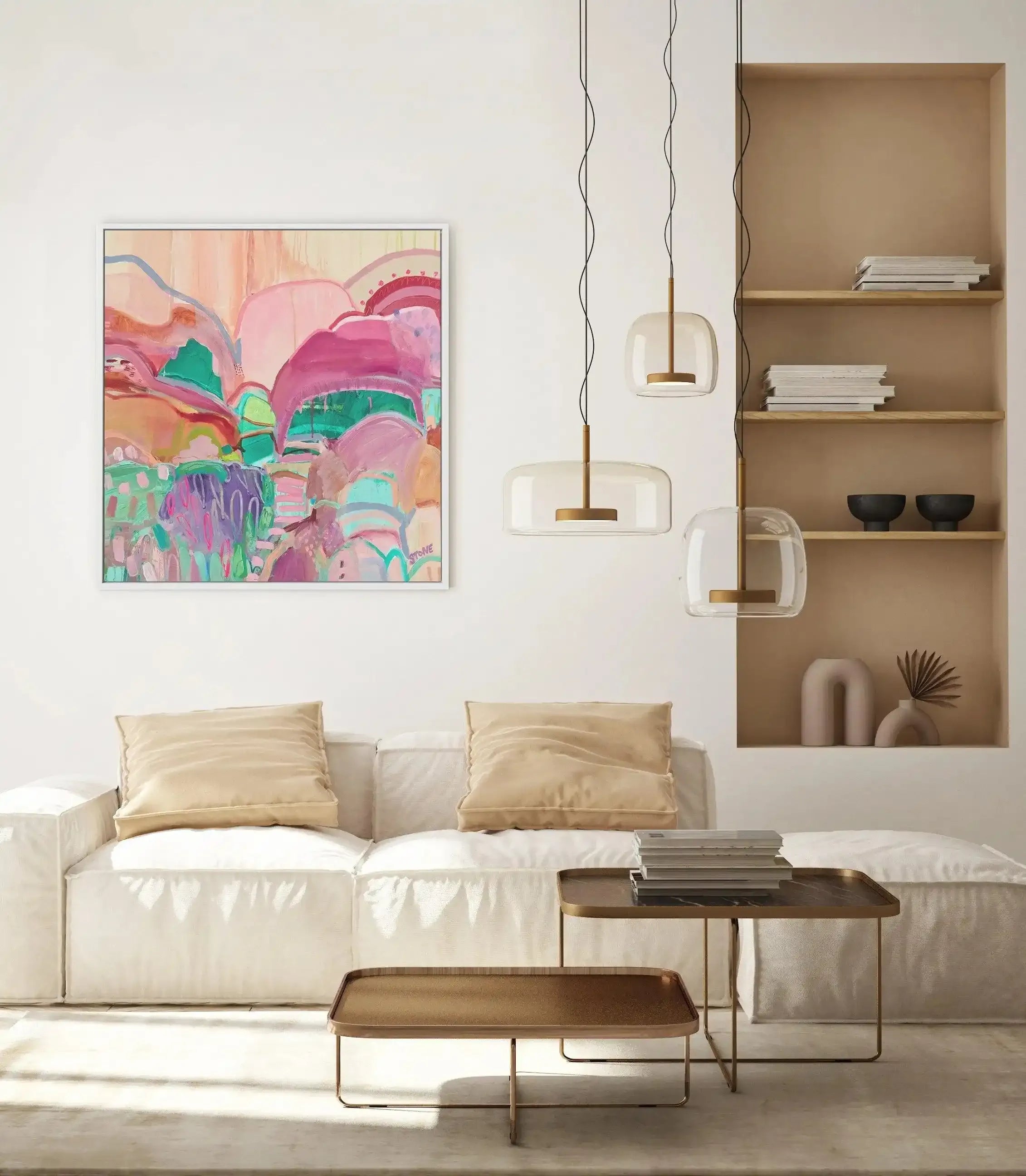 Lifestyle shot of a minimalist living room featuring a large framed abstract art print displayed above a white sofa. The print, titled "Tulum Sunset," features an abstract landscape with rolling hills and organic shapes in shades of pink, fuchsia, teal, and purple, with a white frame and white border. The room includes two light-colored throw pillows on the sofa, two modern gold and glass pendant lights, two gold-framed coffee tables, and a built-in shelf with decorative objects and books.