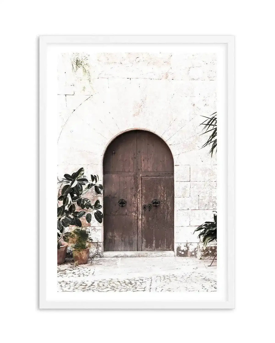 Framed art print featuring a rustic arched wooden door set into a white stone wall, with potted green plants on either side and a cobblestone path in the foreground. The composition has a bright, sun-drenched quality with a natural wood frame and white border.