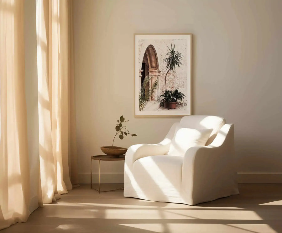 Large framed art print displayed above a curved white armchair in a minimalist living room. The print features a sun-drenched stone archway with potted plants and palm fronds in warm earthy tones against a textured cream wall, with a natural wood frame and white border. The room includes a small round side table with a decorative bowl and a dried plant branch, and sheer cream curtains allowing sunlight to stream in.