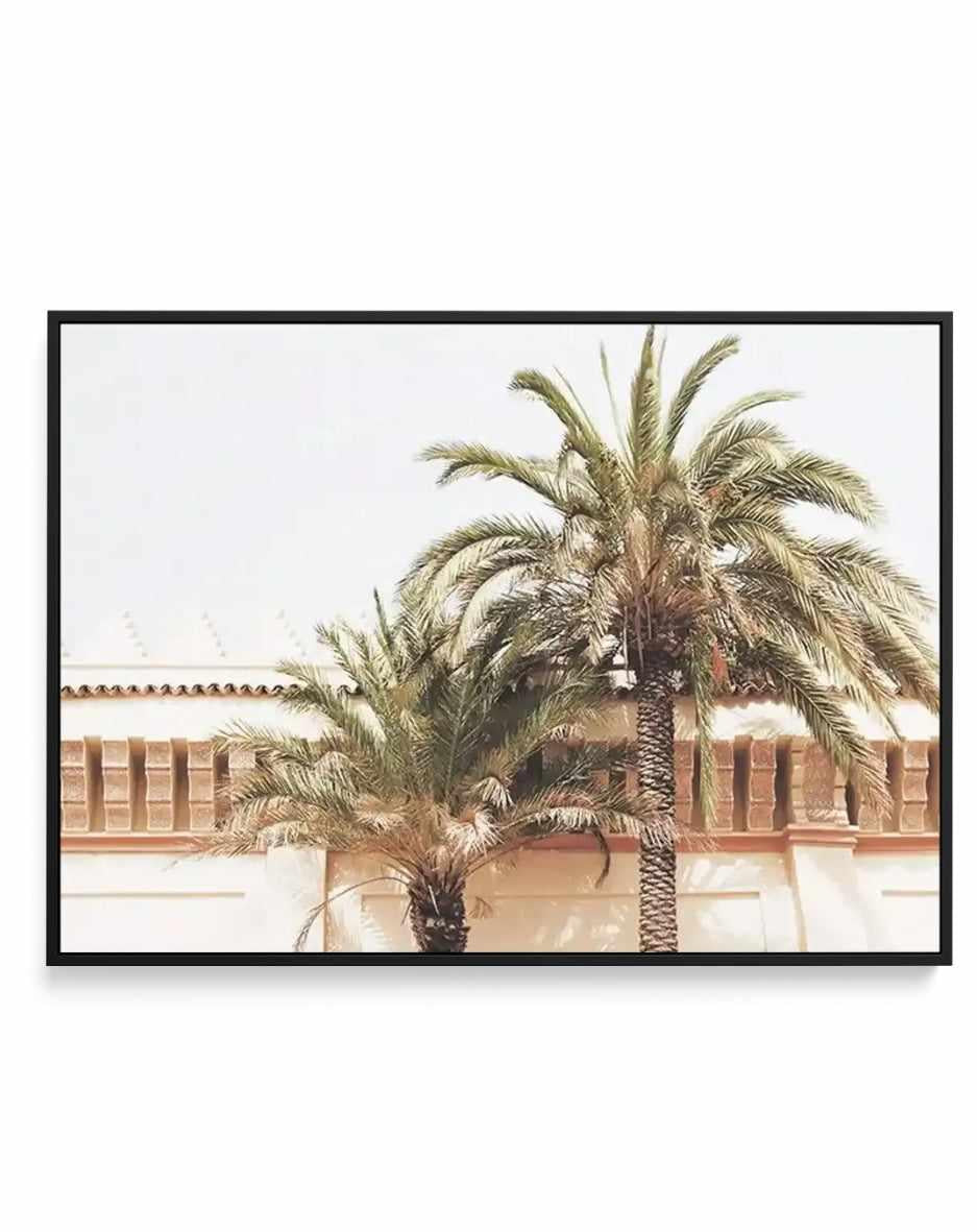 Framed canvas art print featuring two tall date palms with feathery green fronds against a soft, neutral background. A terracotta and cream-colored building with arched windows and decorative tilework is visible behind the palms. The artwork has a muted, warm color palette with dusty greens, tans, and earth tones, rendered in a soft, watercolor-like illustration style. The canvas is mounted in a black frame with a white border.