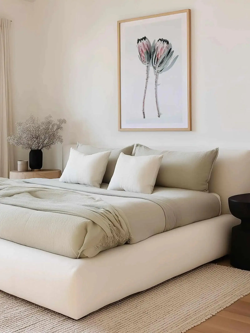 Two minimalist flower illustrations displayed in a natural wood frame with white border, hanging above a cream upholstered bed. The artwork features two delicate protea flowers with dusty rose petals, charcoal-grey leaves, and thin grey stems rendered in a loose watercolor style against a white background. The bedroom setting shows neutral tones with cream and beige bedding, a wooden nightstand with a black vase of dried baby's breath, and a natural fiber area rug, creating a soft, botanical aesthetic.