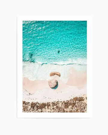Aerial photography art print of a beach scene with vibrant turquoise ocean meeting a white sandy shore. The composition features two large rocks on the sand, one partially submerged in a shallow pool of water, and a line of brown coastal vegetation at the bottom.
