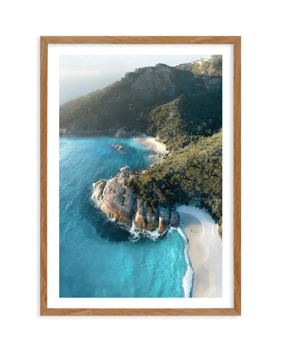 Framed aerial photograph of a coastal scene with a natural wood frame and white border. The image captures clear turquoise water meeting a white sand beach, with a large rocky outcrop covered in green foliage separating two coves. Lush green hills rise behind the beaches under a bright, slightly hazy sky.