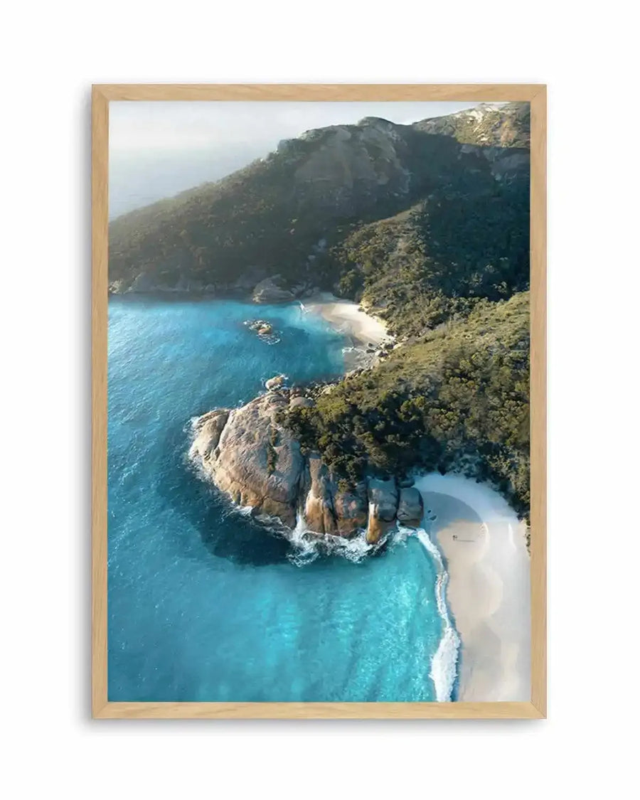 Framed aerial photograph of a coastal scene with vibrant turquoise water, white sandy beaches, and lush green hills. The composition features a large rocky outcrop dividing two crescent-shaped beaches, with gentle waves breaking on the shore, displayed in a natural oak frame with white border.