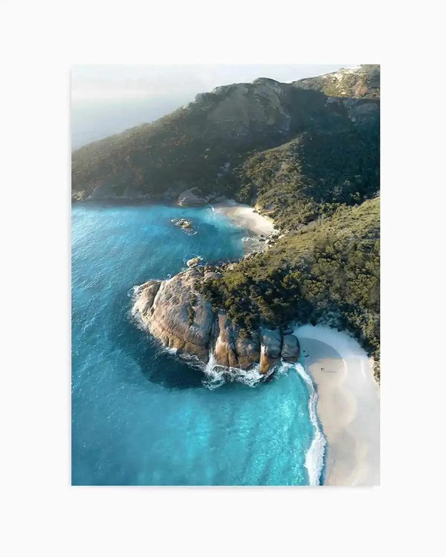 "Aerial photograph art print of a coastal bay with turquoise water, white sand beaches, and lush green hills. The composition features a large rocky outcrop in the foreground with waves crashing against it, and two smaller beaches nestled between the hills and the clear blue ocean."