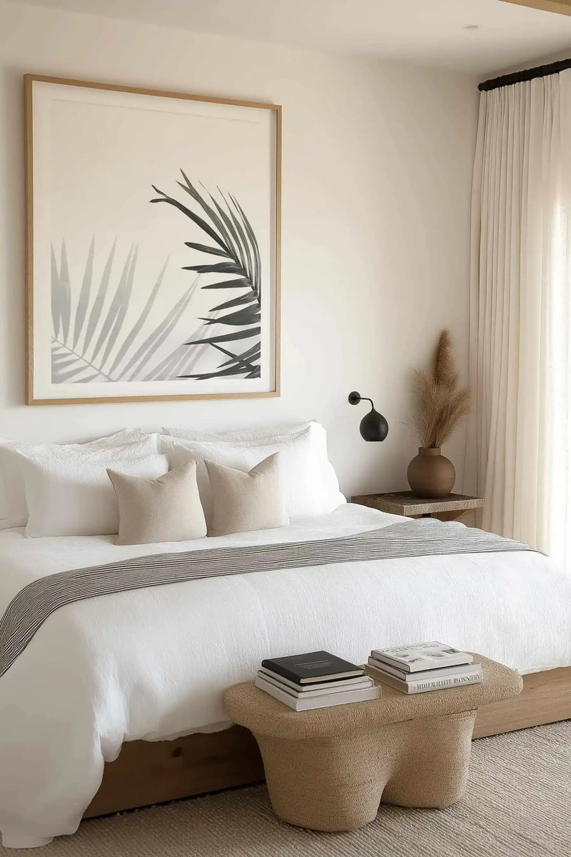 Framed art print displayed above a bed showing an illustration of palm fronds in shades of gray and black against a cream background. The print has a natural wood frame with a white border. The bedroom features white bedding, cream throw pillows, a striped gray bed runner, cream curtains, and a woven bench at the foot of the bed holding stacked books and a decorative vase with dried pampas grass.