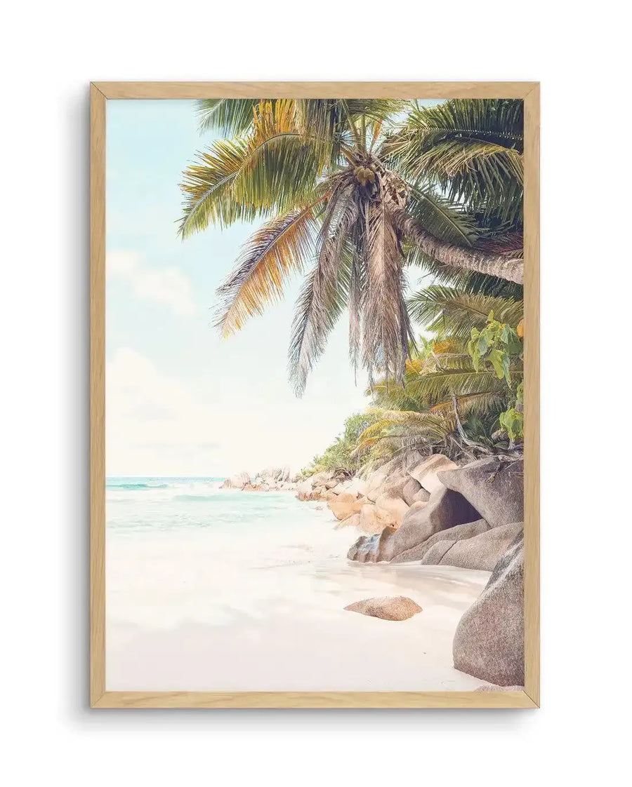 Framed photography art print of a tropical beach scene in Seychelles, with a natural wood frame and white border. The composition features a white sand beach with turquoise ocean and gentle waves on the left, and large granite boulders and lush green palm trees on the right, under a soft blue sky.