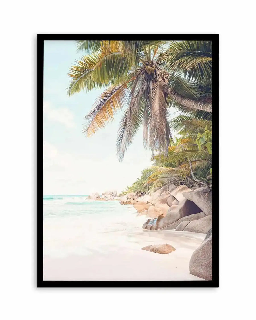 Framed photography art print of a tropical beach scene in Seychelles with a black frame and white border. The composition features a palm tree with green and brown fronds hanging over a white sandy beach, with turquoise ocean waves gently lapping the shore and large grey boulders scattered along the coastline under a clear, pale blue sky.