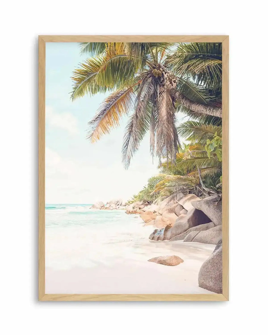 Framed photography art print of a tropical beach scene with a natural wood frame and white border. The composition features a palm tree with green and brown fronds hanging over a white sandy beach with turquoise ocean water and large light brown rocks along the shore under a pale blue sky.
