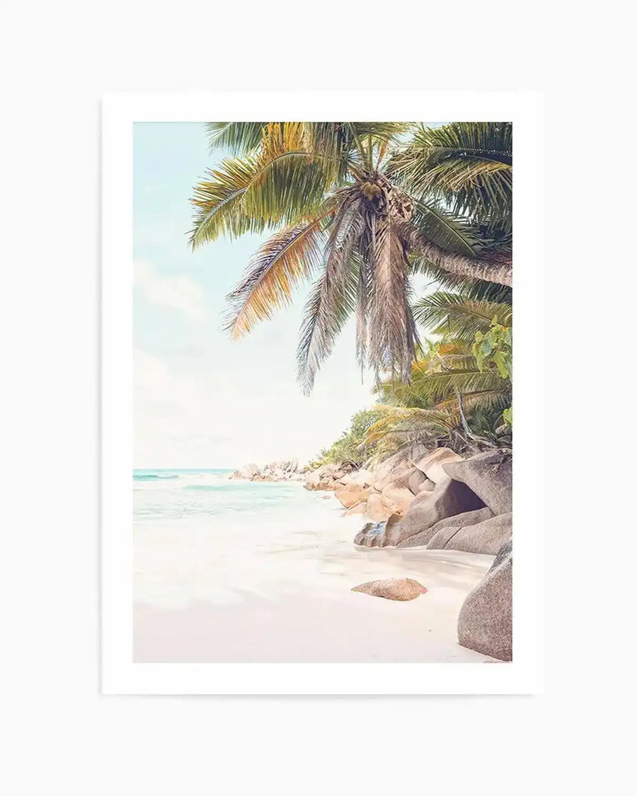 Framed photography art print of a serene tropical beach scene with a natural wood frame and white border. The composition features a large palm tree with green and dried fronds arching over a white sand beach, with turquoise ocean waves gently lapping the shore and large smooth boulders scattered along the coastline under a pale blue sky.